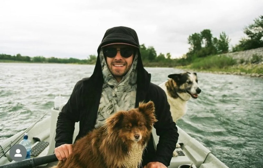 Person in a boat on a river with two dogs, wearing sunglasses and a hooded jacket.