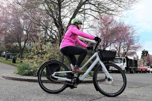 Person in pink jacket and green helmet riding a bicycle on a street with blooming trees.