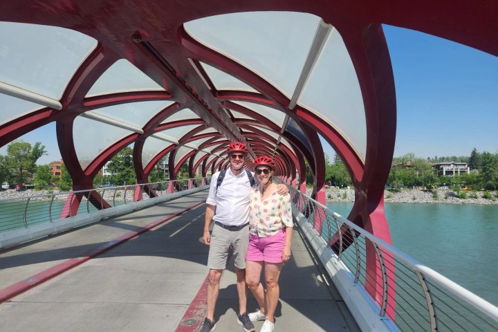 Two people with helmets stand smiling on a red arched bridge over a river.