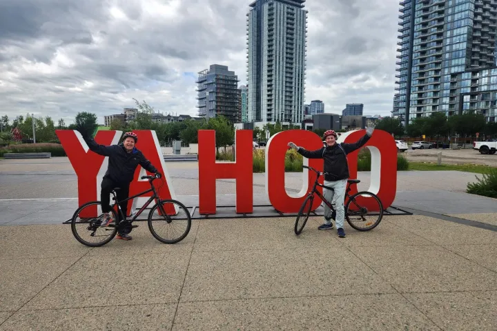 Two cyclists pose by large red 'YAHOO' letters in front of city skyscrapers.