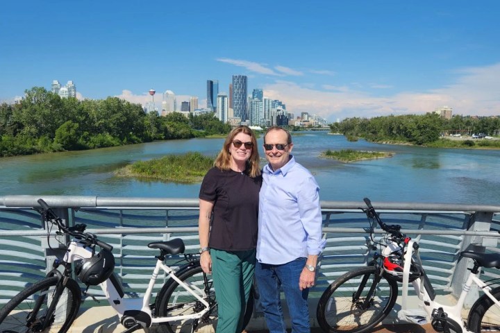 Couple posing with bicycles on a bridge, city skyline and river in the background.