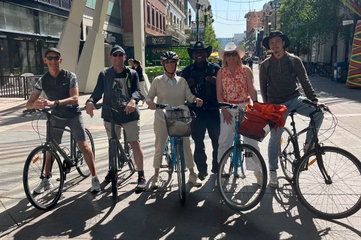 Six people with bikes posing on a sunny urban street with tall buildings.