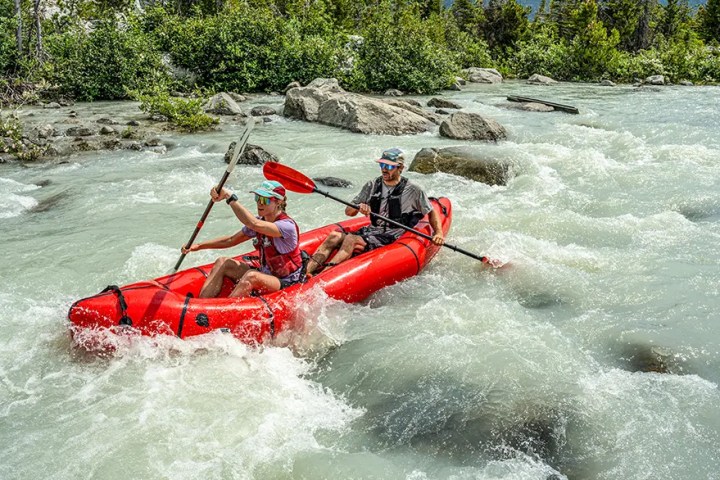Two people paddling a red inflatable kayak in a rapid river surrounded by greenery.