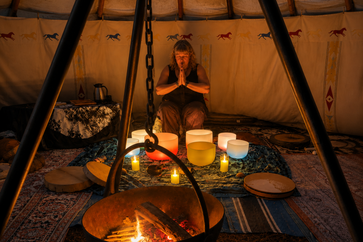 Person meditating with sound bowls and candles in a tipi with a fire pit in the foreground.