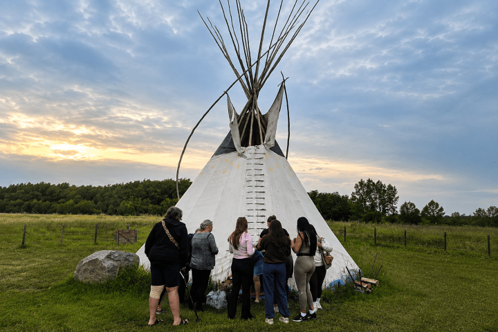 Group of people outside a teepee on a grassy field at sunset.
