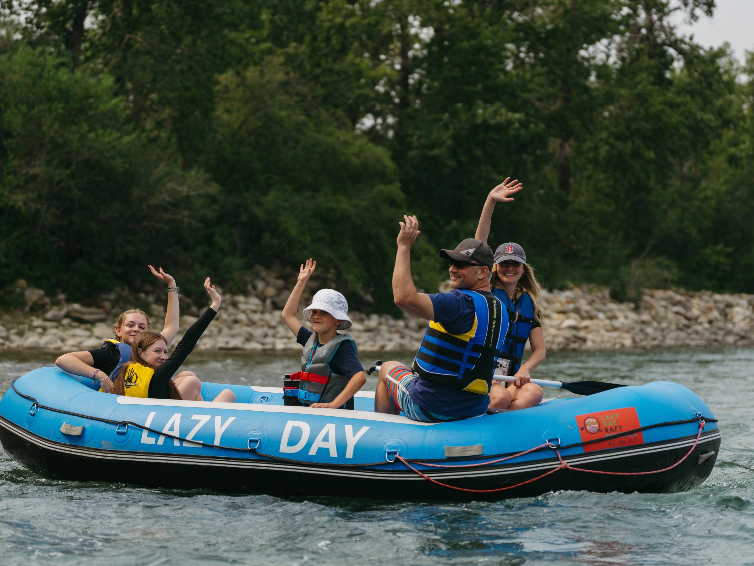 Family waving while rafting on a blue inflatable raft named 'Lazy Day' on a river.