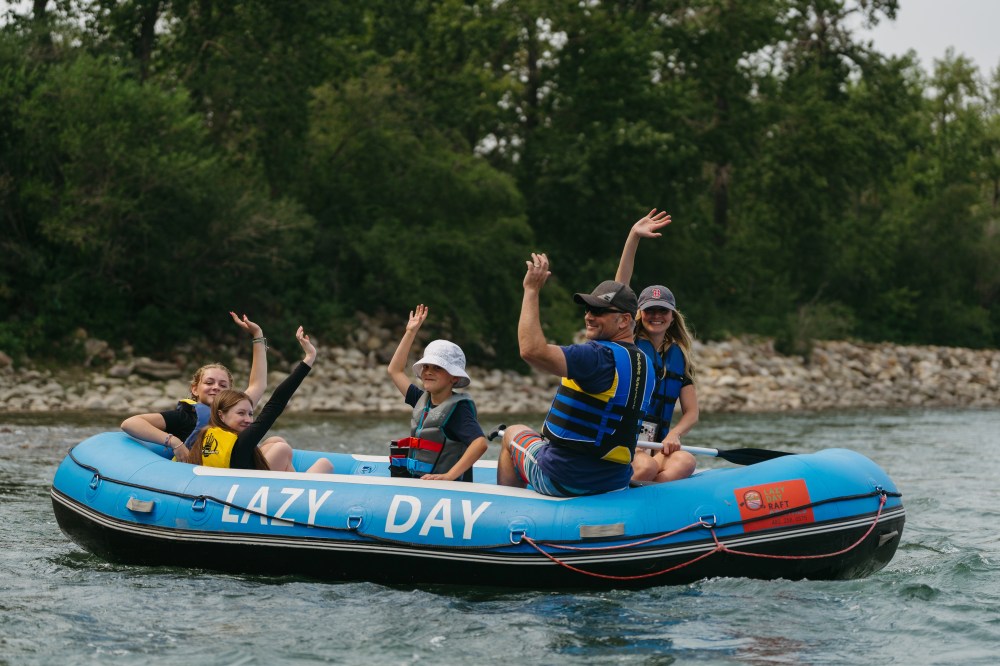 Family waving while rafting on a blue inflatable raft named 'Lazy Day' on a river.