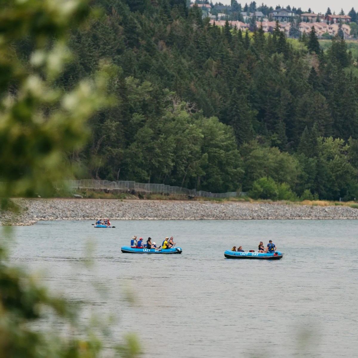 People rafting on a river surrounded by trees and distant buildings.