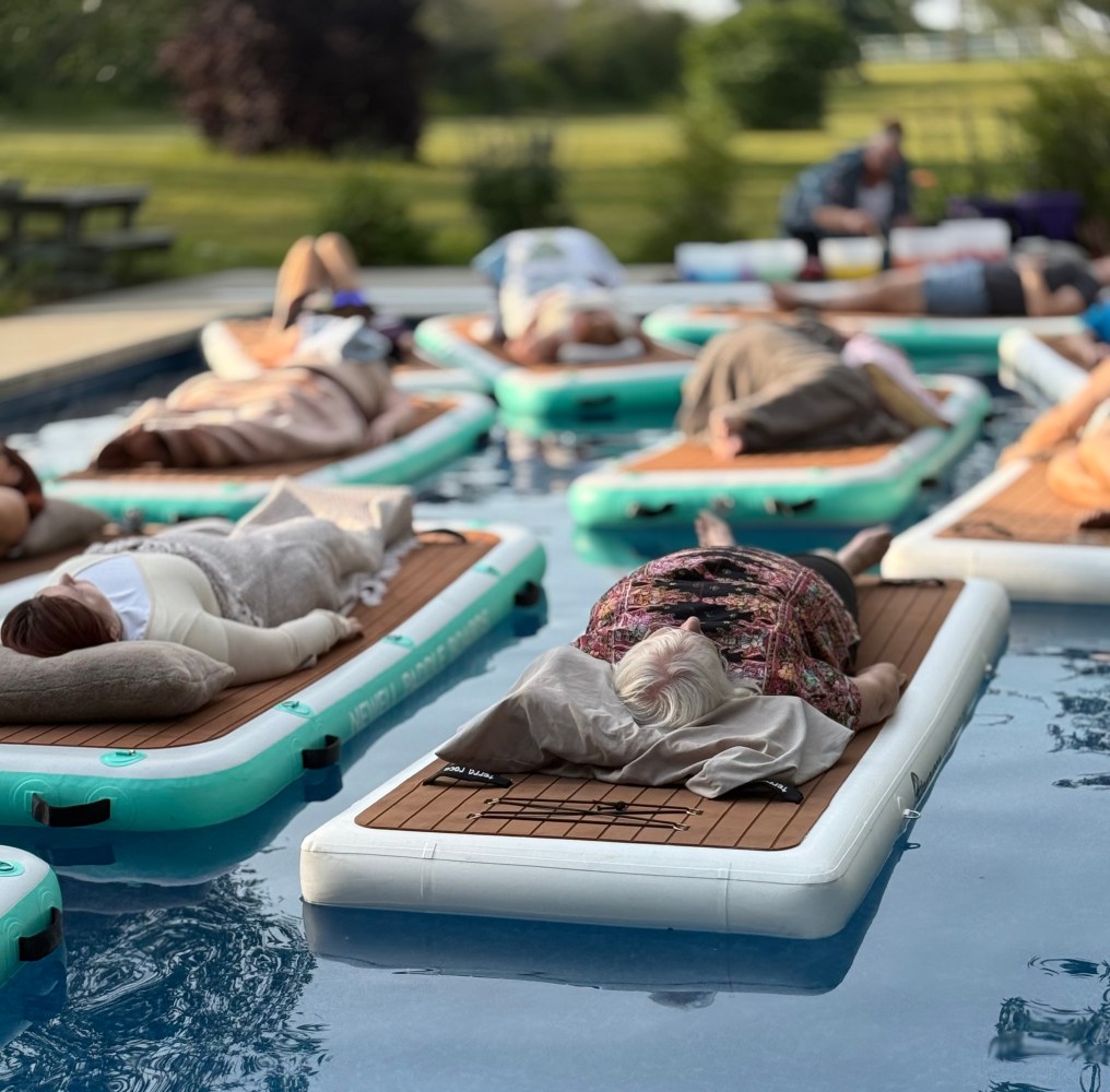 People relaxing on floating mats in a pool with greenery in the background.