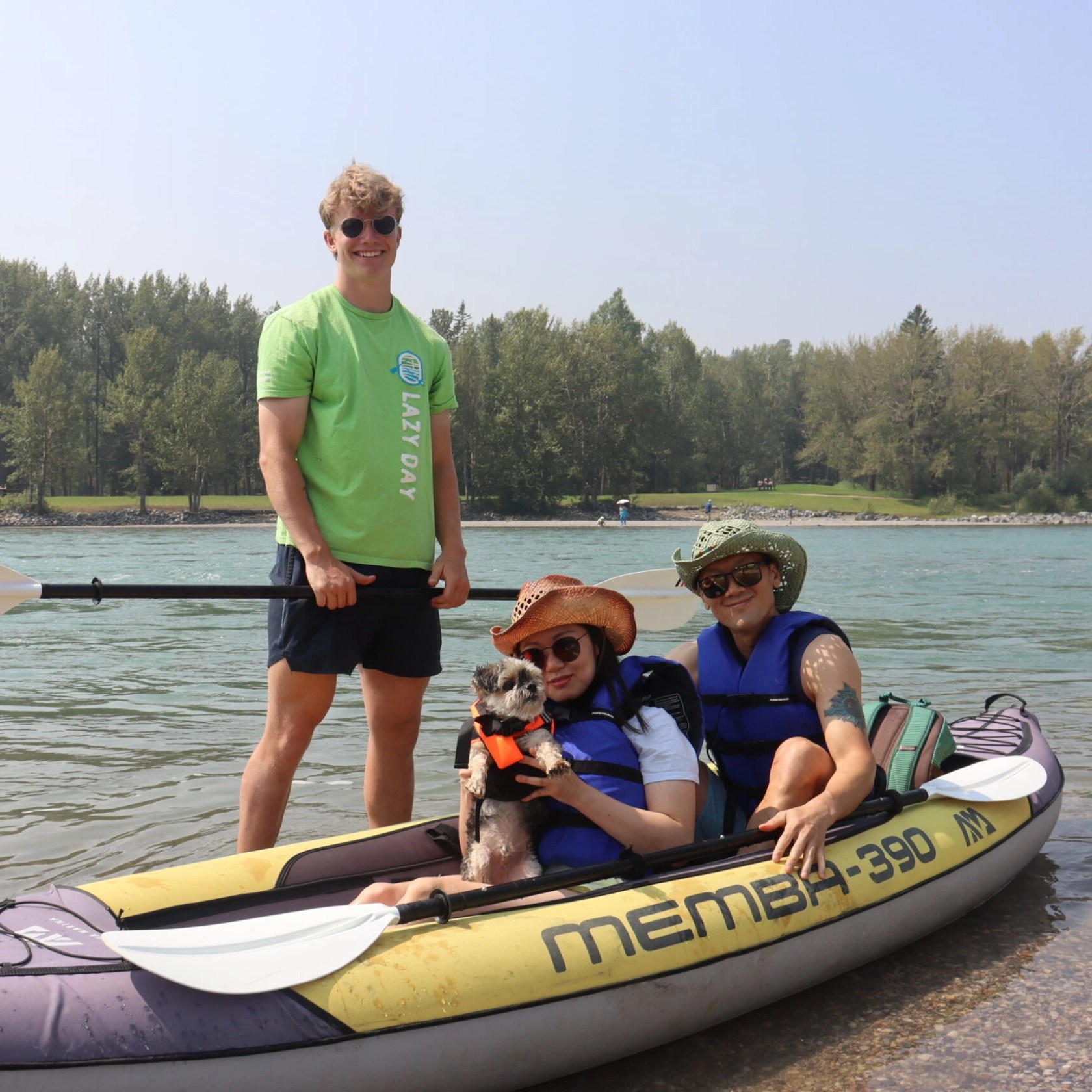Three people and a dog in a kayak by a river, with one person standing holding a paddle.
