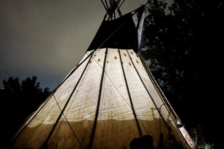 Illuminated tipi at night with trees in the background.