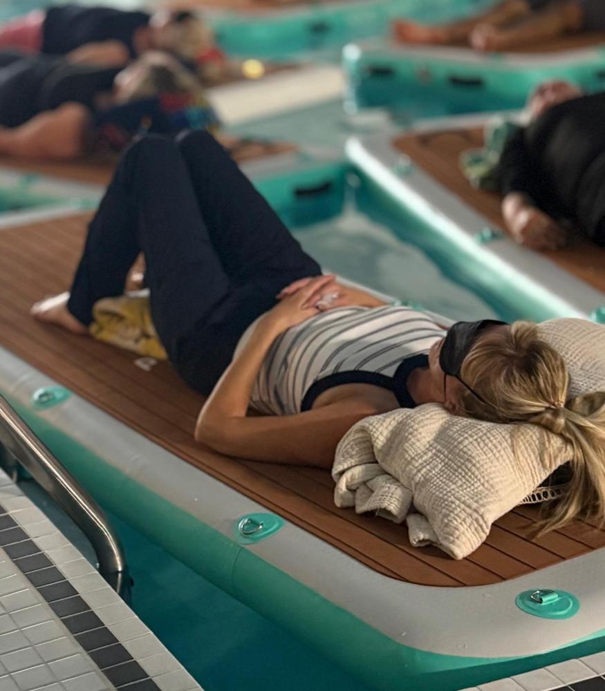 Person resting on a floating board in a pool, using pillows and wearing an eye mask.