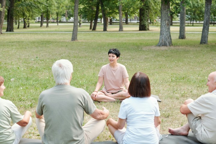 Group of people sitting on grass in a park, listening to a speaker.