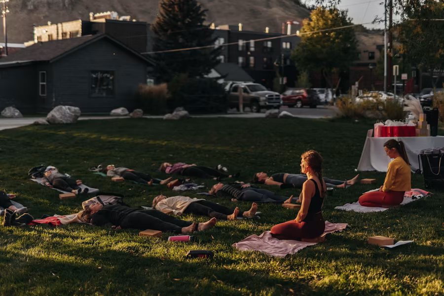 People lying on grass in yoga session outside, one person sitting up near them.