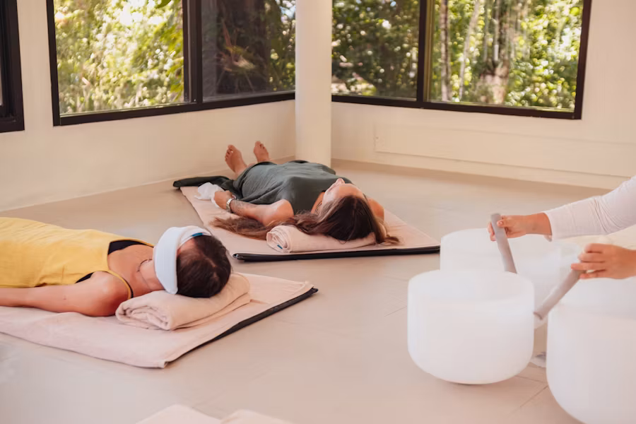 Two people lying on mats indoors, wearing eye masks, with sound bowls nearby.