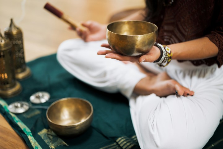 Person holding a brass bowl, seated cross-legged on a mat with prayer items.