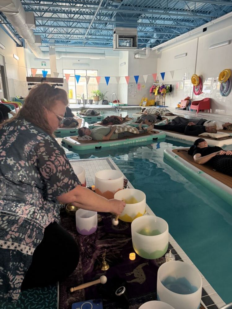 Person playing singing bowls by a pool; people relax on floating mats.
